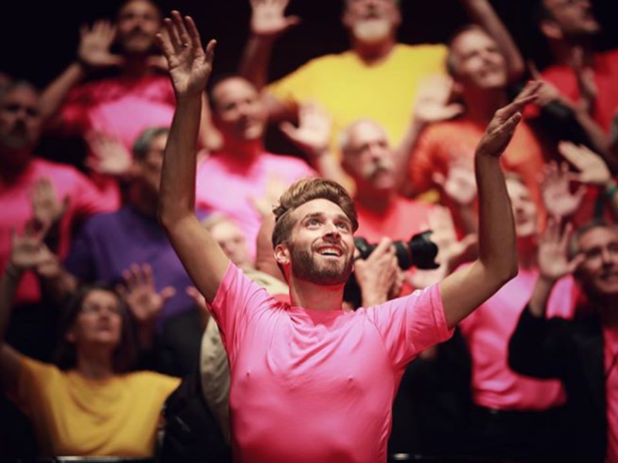 A man wearing a pink shirt posing while dancing, arms raised and looking upward