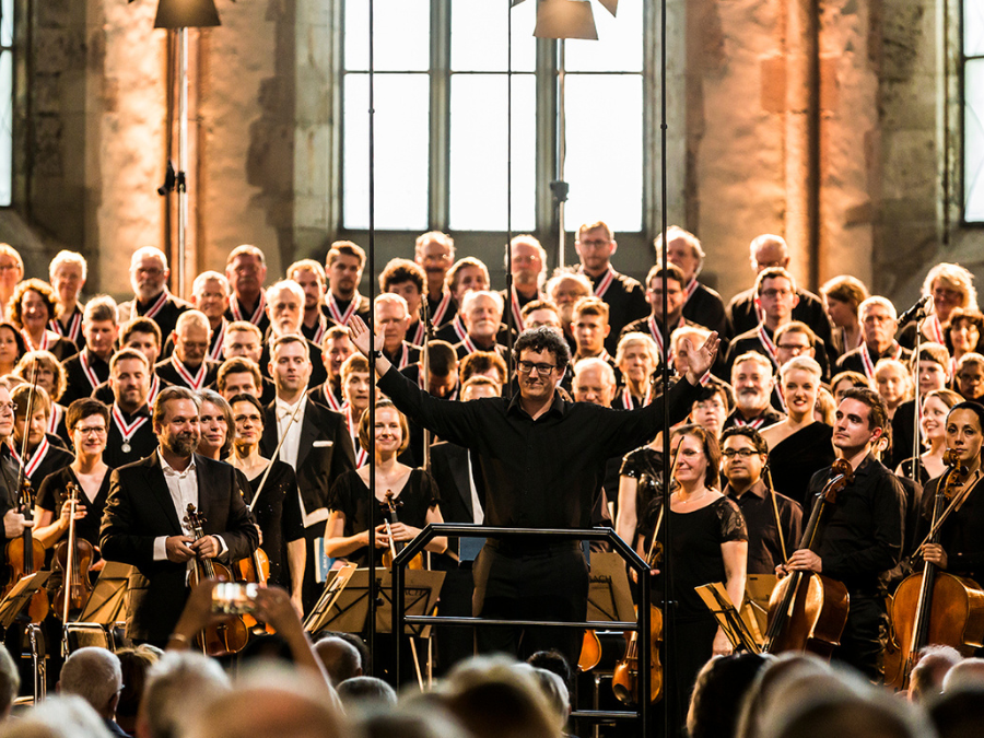 A choral conductor standing in front of a choir and orchestra with his arms raised as the audience applauds