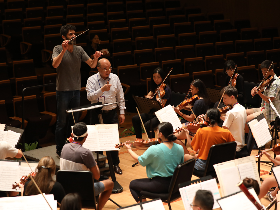 A conductor leading an orchestra in rehearsal while a composer listens