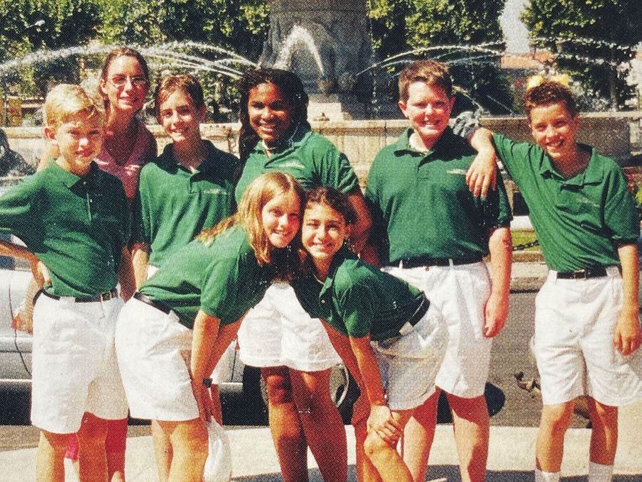 Smiling kids dressed in green polos and white shorts standing in front of a fountain